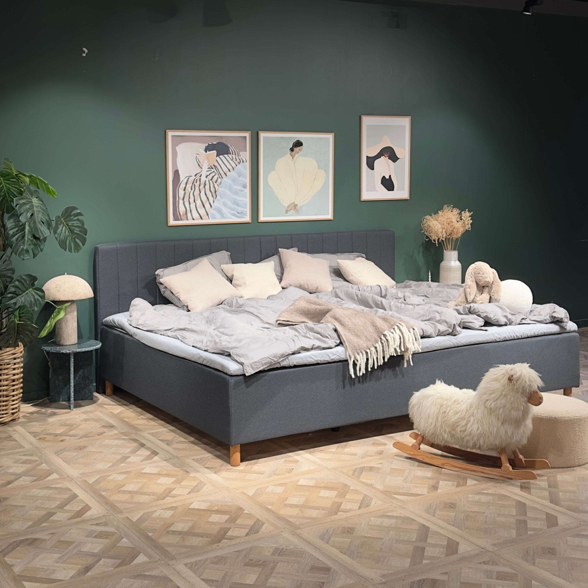 Cama familiar larga, cinzenta e feita, num quarto com uma parede verde e uma ovelha de baloiço no chão