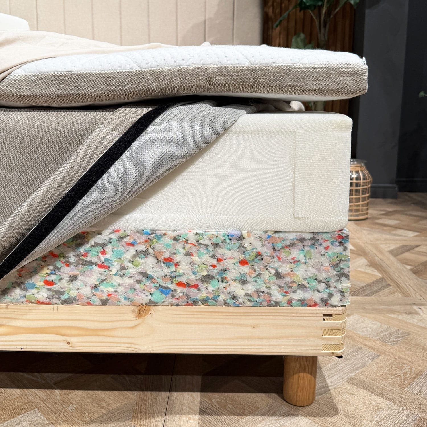 Camadas da cama abertas num dos cantos: sobrecolchão, colchão de molas com a capa aberta e rebordo em espuma da base da cama sobre a estrutura de madeira