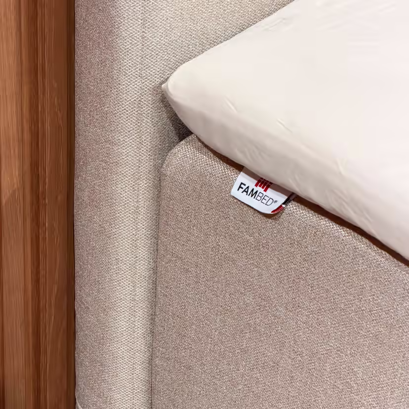 Grande plano da capa bege que envolve o canto da cama, com a etiqueta FAMBED e um lençol branco por cima