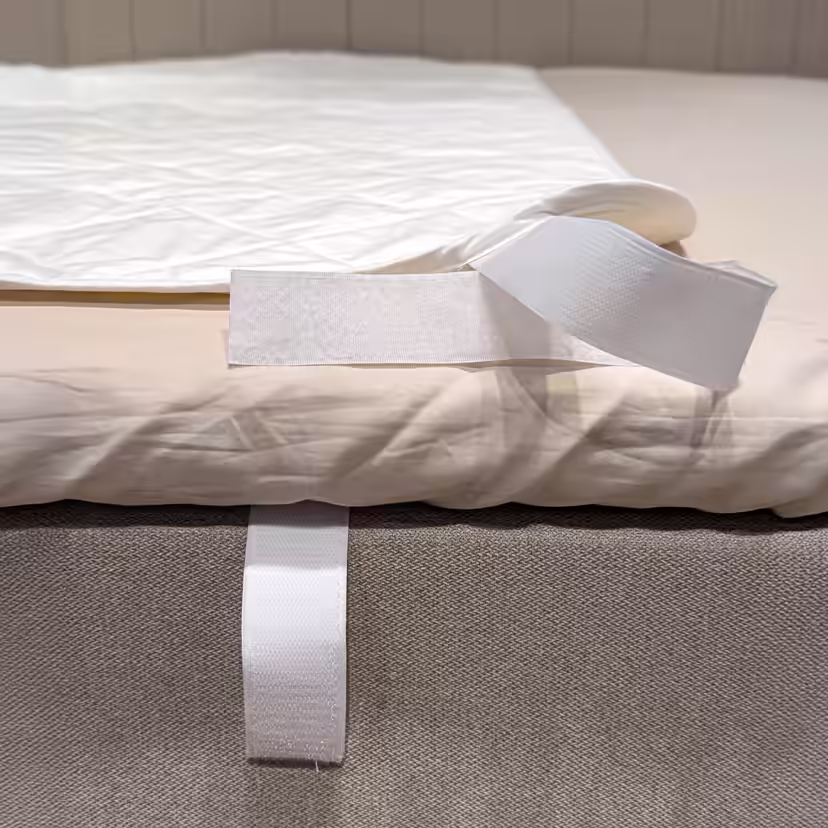 Canto do protetor de colchão branco e acolchoado dobrado para trás, deixando visível a tira de velcro à volta do rebordo da cama