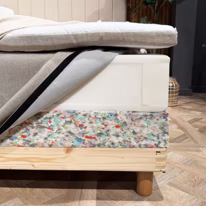 Camadas da cama abertas num dos cantos: sobrecolchão, colchão de molas com a capa aberta e rebordo em espuma da base da cama sobre a estrutura de madeira